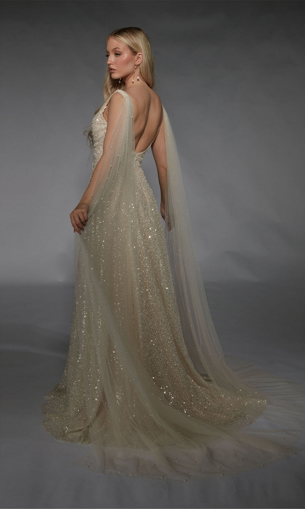 A woman poses in the Long White Dress: Alyce 7134, a floor-length evening gown with sequin embellishments, low back, and sheer cape sleeves, set against a gray backdrop.